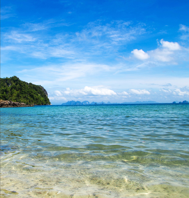 Shallow water leading out into an ocean. You can see the sand under the water, blue sky with white puffy clouds and sunshine in the background.
